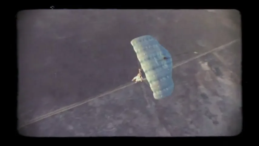 A person with a light blue parachute descends over a vast, arid landscape with faint roads, filmed from an aerial perspective.