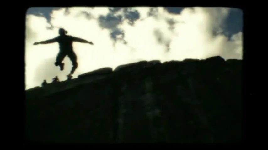 A silhouetted person in a hat and casual clothes jumps off a cliff against a bright sky with clouds, captured in a vintage film style.