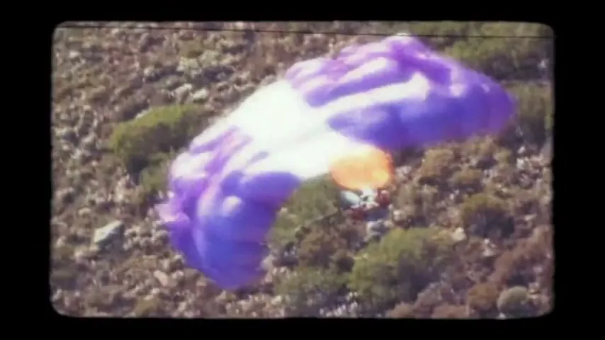 A person in a blue jumpsuit with a purple and orange parachute is seen from above, descending over a rocky, scrub-covered hillside on a sunny day.