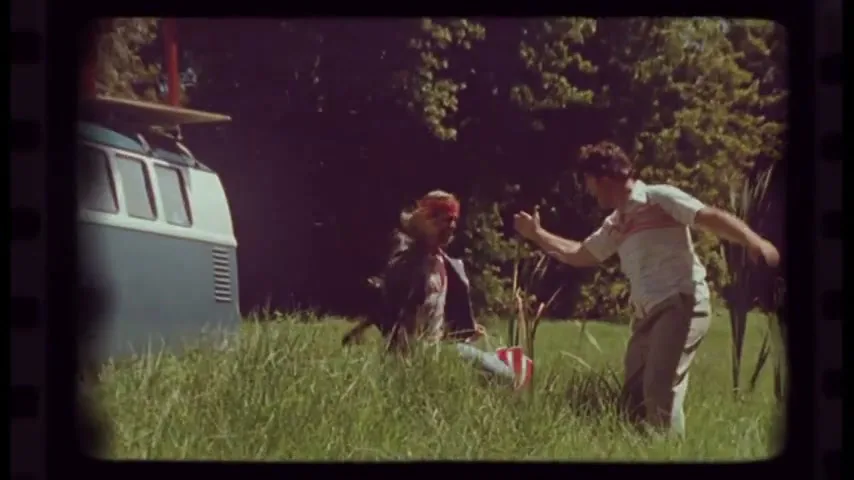 Two men in a grassy field struggle over a parachute next to a blue and white van on a sunny day.