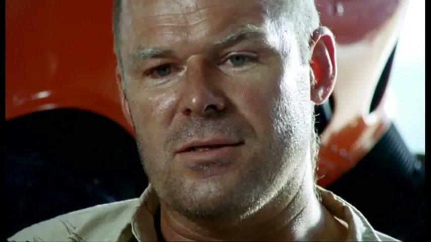 A man with a short beard and light shirt looks concerned, with an orange plane visible in the background.
