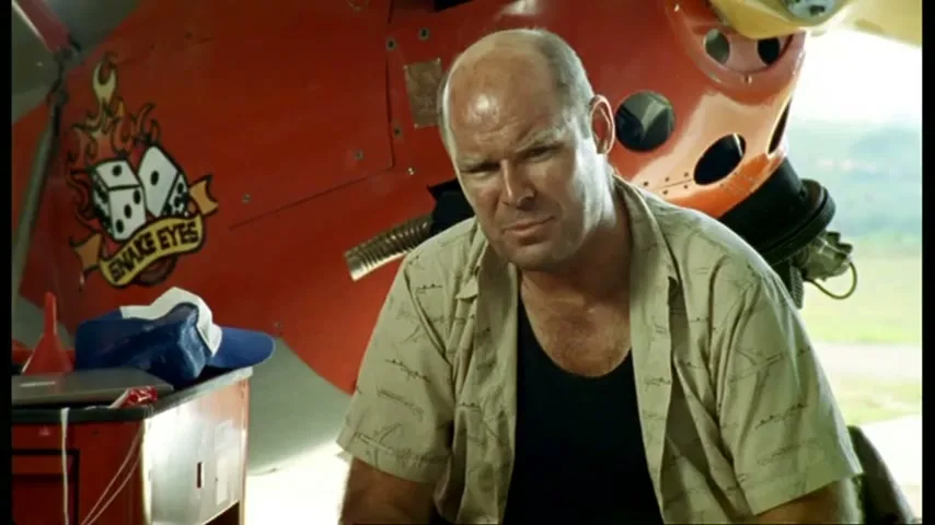 A bald man in a patterned shirt and black undershirt talks to the camera, sitting in front of a red plane with a "Snake Eyes" logo.
