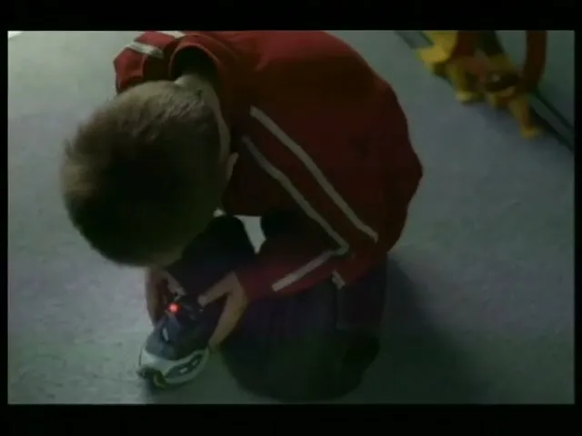 A young boy in a red top and dark trousers kneels on a grey floor, looking down at his blue and white shoe with a red light on it.
