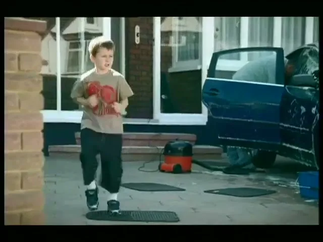 A young boy in a t-shirt and shorts runs in place on a paved driveway in front of a house, while a blue car is being washed in the background.
