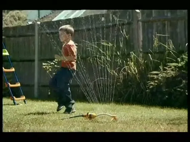 A young boy in a red shirt and blue jeans jumps through a sprinkler in a sunny backyard.