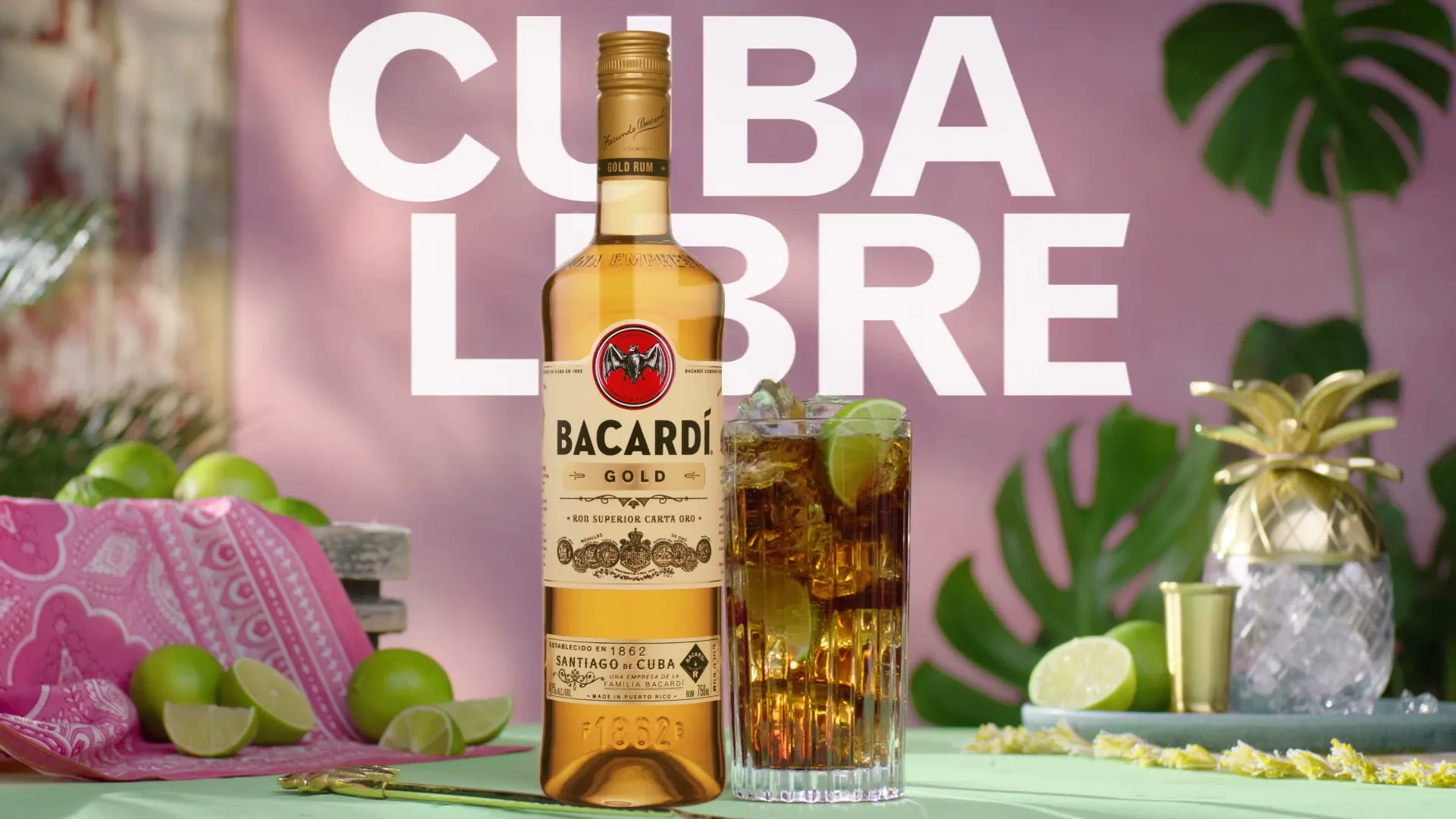 A bottle of Bacardi Gold rum and a glass of Cuba Libre cocktail are displayed on a table with limes and tropical leaves.