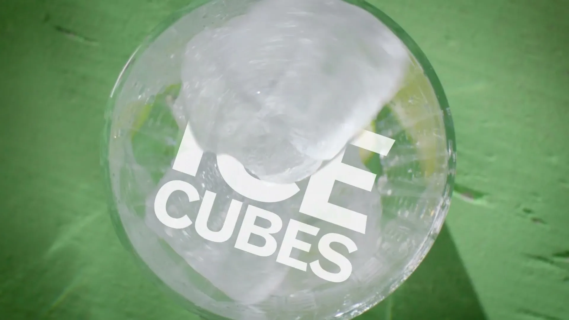 Ice cubes and lime wedges are dropped into a glass, seen from directly above, on a green surface.