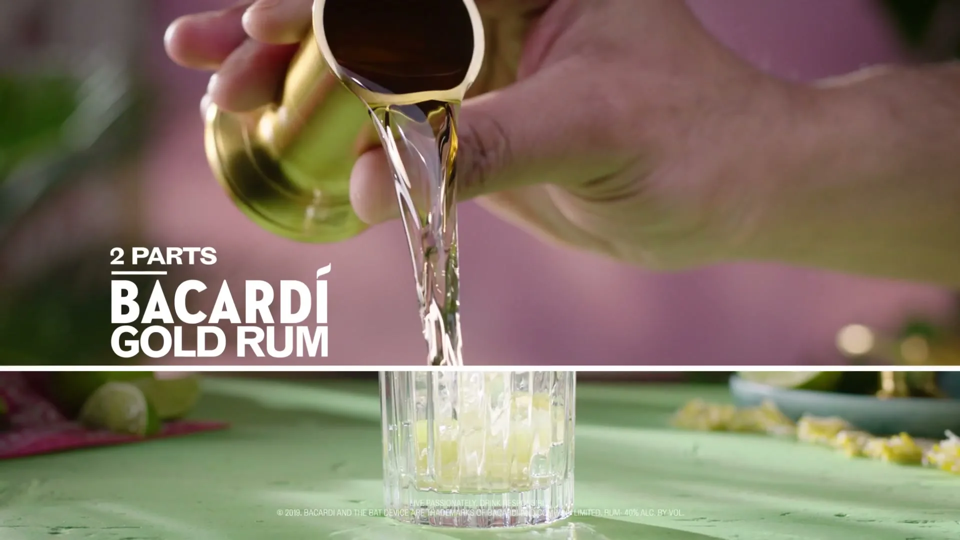 A bartender's hand pours Bacardi Gold Rum from a jigger into a glass with lime wedges on a green surface.