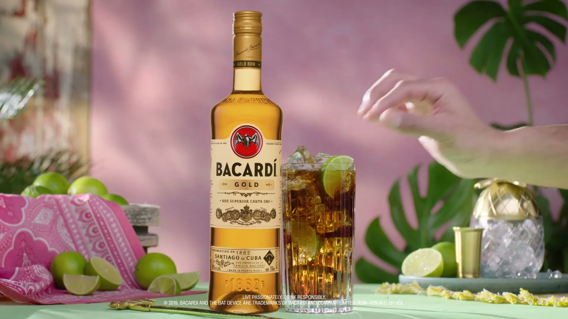 A hand places a lime wedge into a tall glass of Bacardi Gold rum and cola, surrounded by limes and tropical decor on a table.