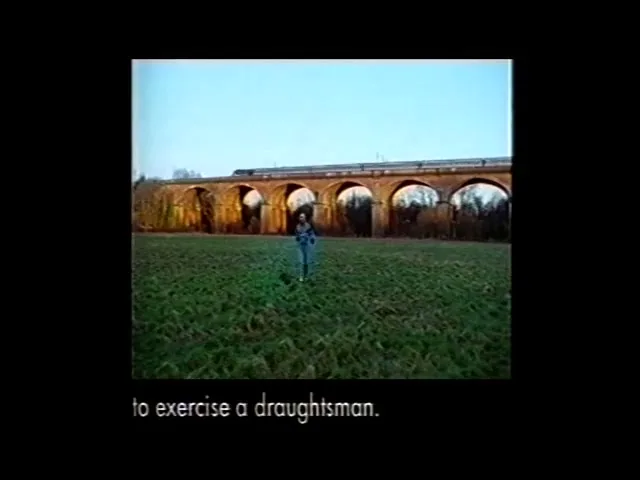 A young man in a dark jacket and light pants walks across a grassy field towards a brick viaduct with a train on top, under a clear sky.