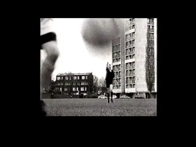 A child in a striped shirt kicks a soccer ball in a grassy field, with apartment buildings in the background under an overcast sky.