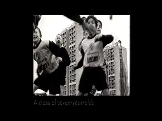 A man in a dark jacket and shorts stands in front of a tall building, directing children who run past him in a black and white, grainy shot.