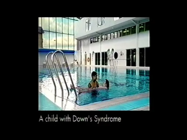 A female swimming instructor in a black swimsuit helps a child with Down syndrome float on her back in a bright indoor swimming pool.