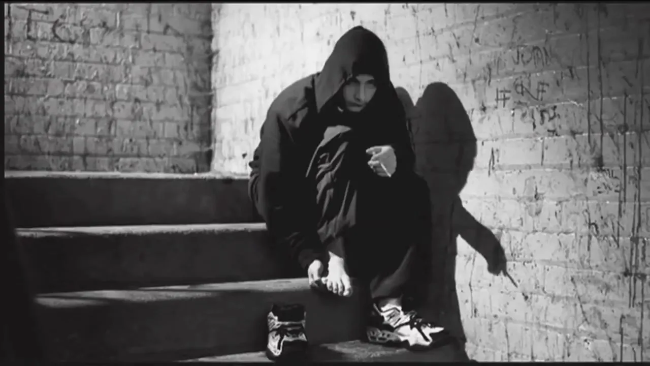 A young person in a dark hoodie sits barefoot on concrete steps, holding a cigarette, in a gritty, graffiti-covered urban stairwell.
