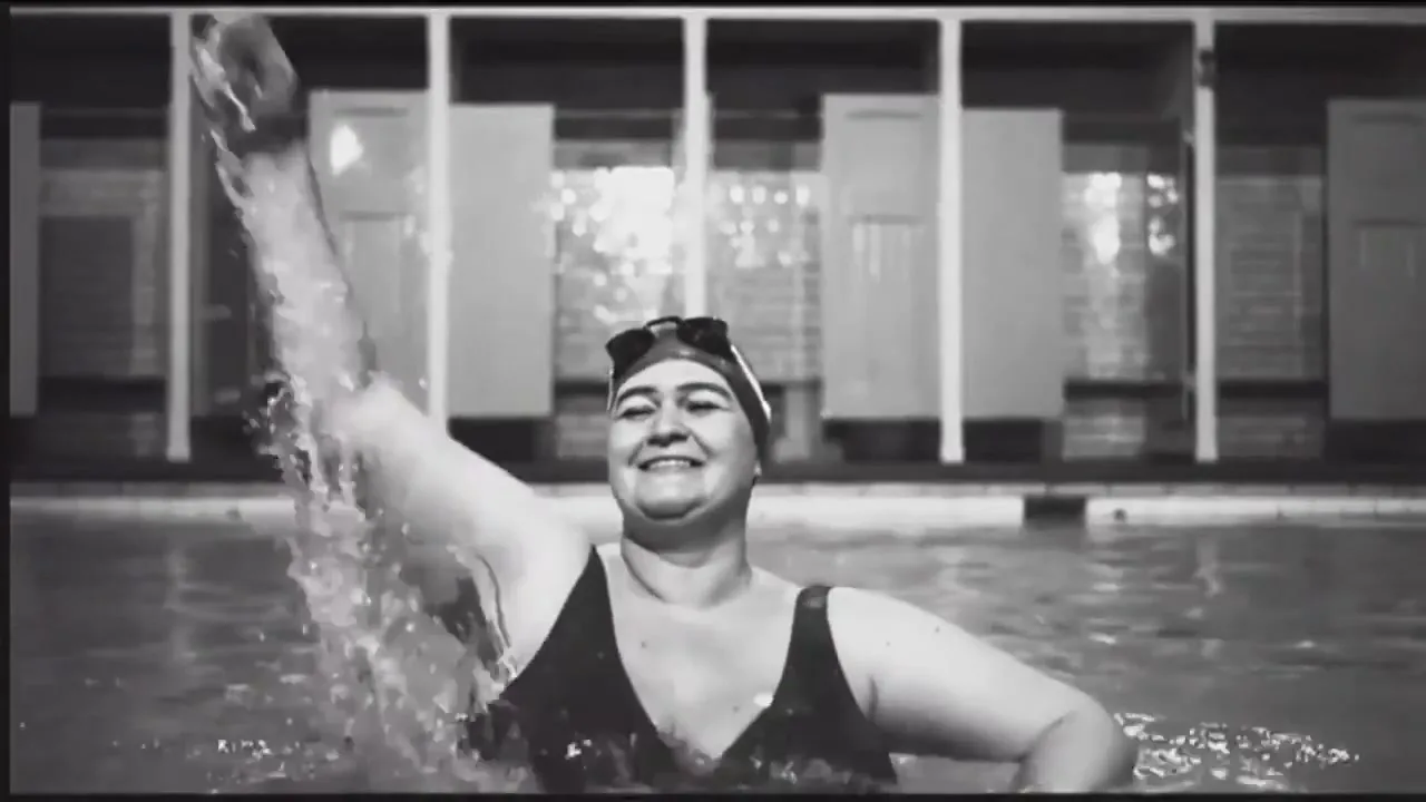 A smiling woman in a swimming cap and goggles stands in a pool, raising her arm and splashing water in an indoor swimming pool.