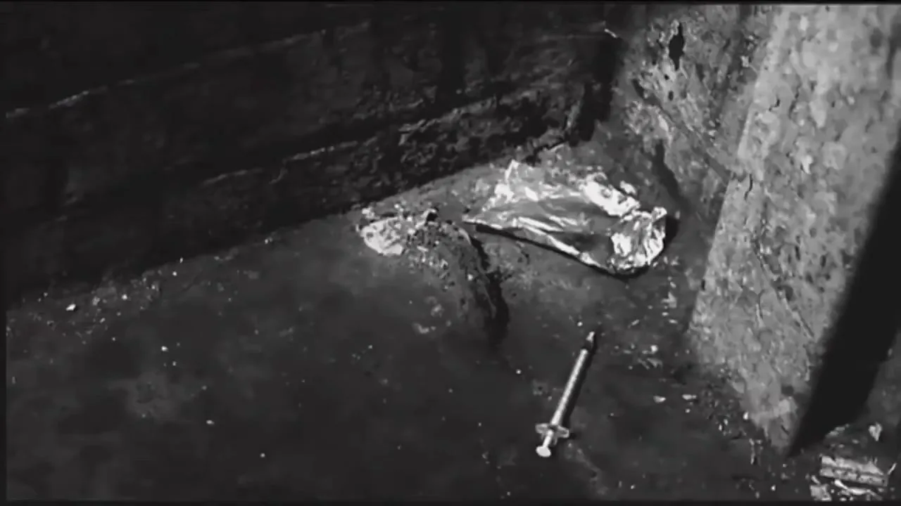 A used syringe and crumpled foil lie on a dirty, dark floor in a close-up shot, suggesting a grim, neglected environment.