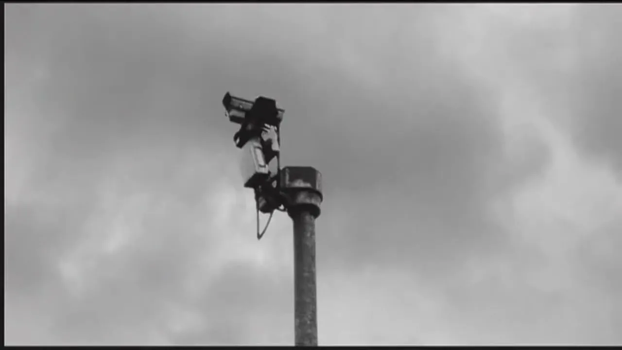 A black and white shot of a security camera on a pole against a cloudy sky, with a whisper heard.