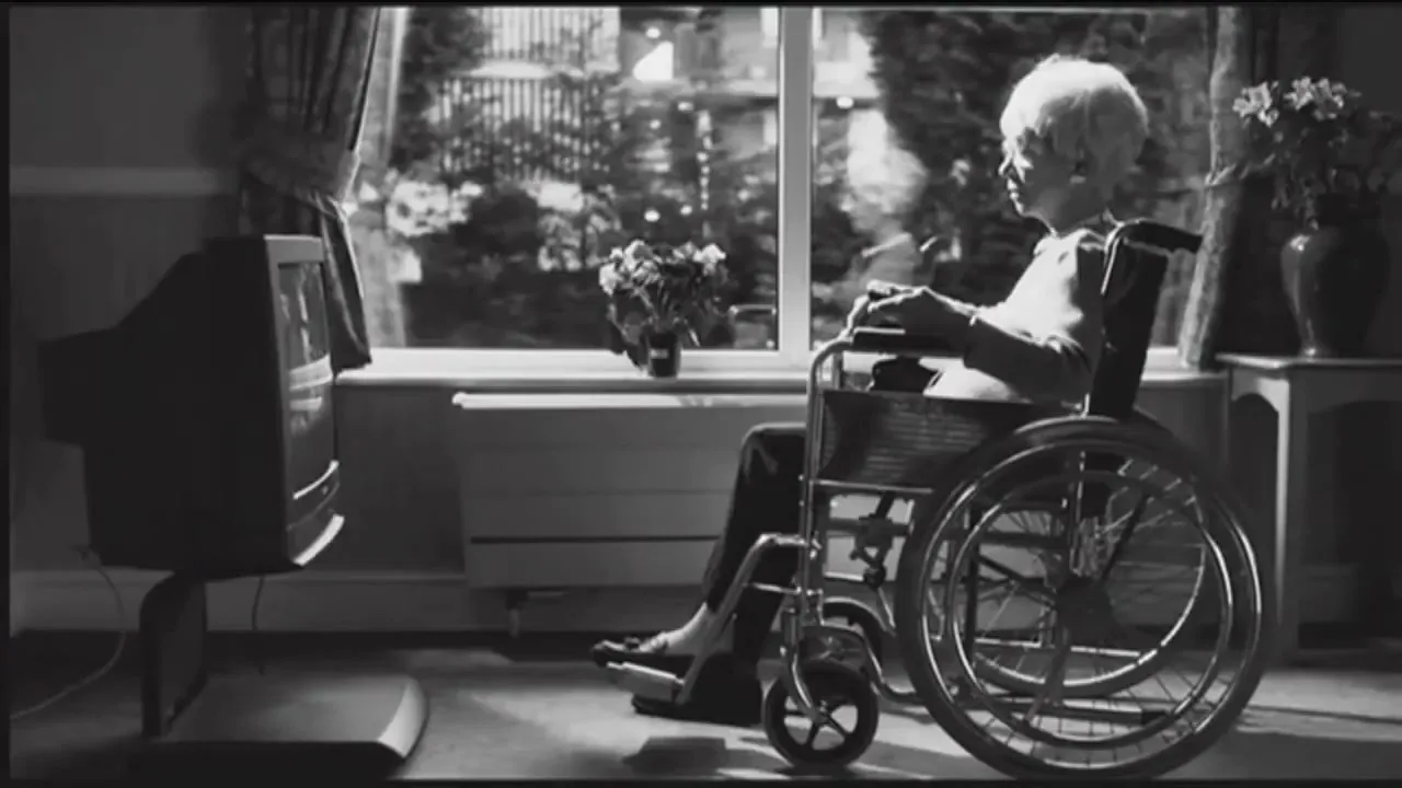 An elderly woman with white hair sits in a wheelchair, looking out a window in a dimly lit room, with a television set to her left.