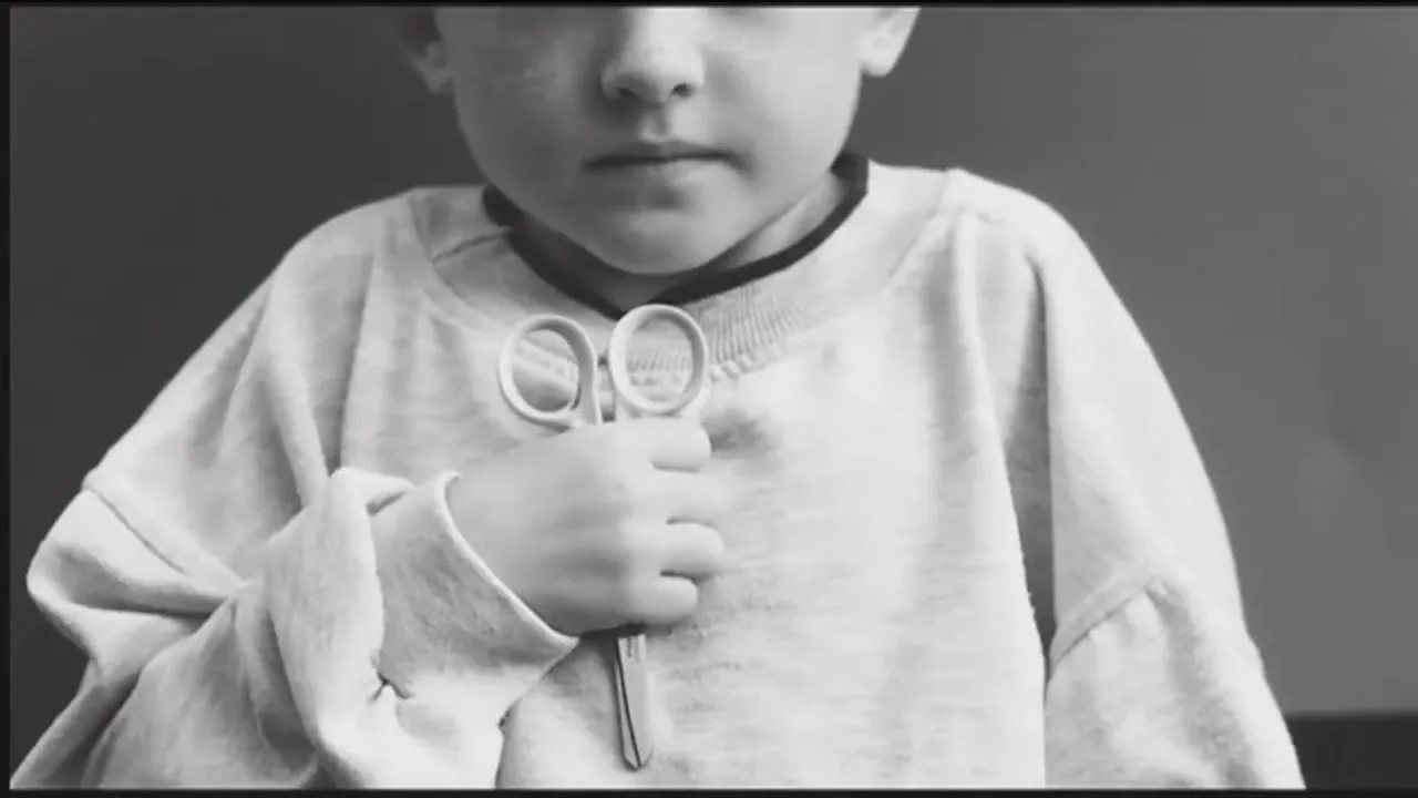A young child, dressed in an oversized light-colored sweatshirt, holds a pair of scissors in their hand, looking directly at the camera in a medium shot.