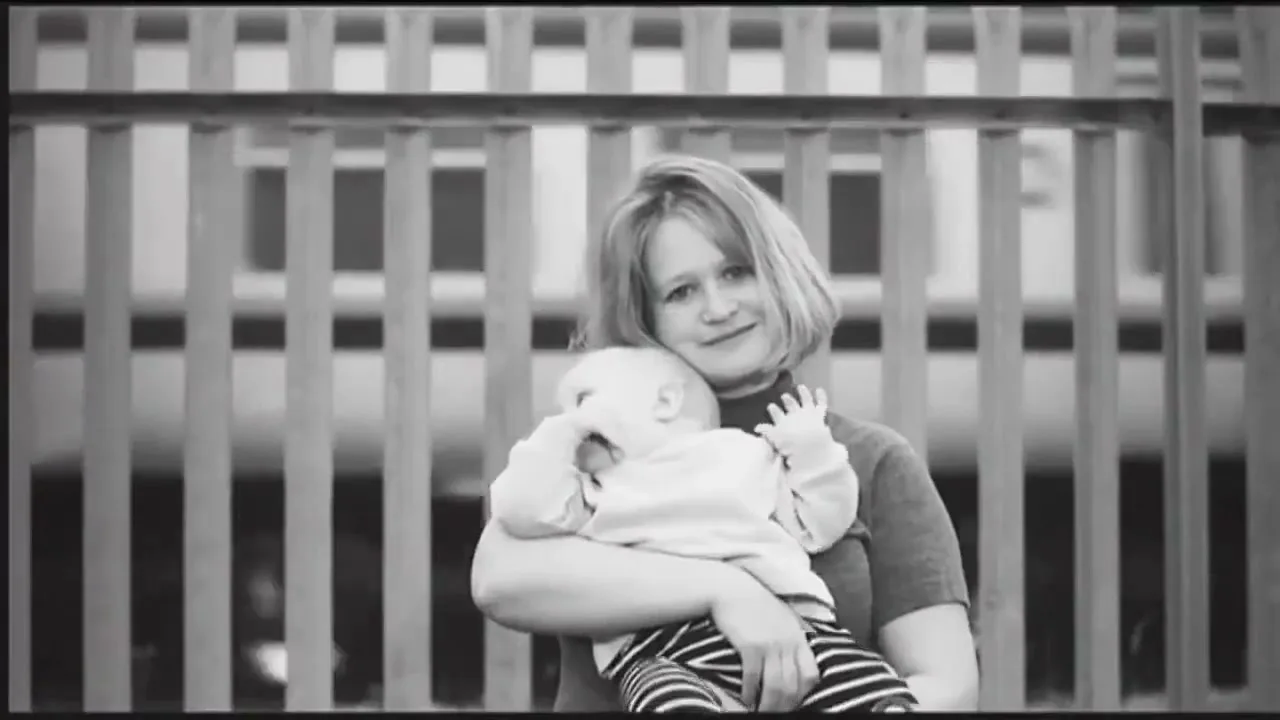 A woman with short hair holds a baby in her arms, both looking at the camera with a gentle smile, in front of a blurred fence.