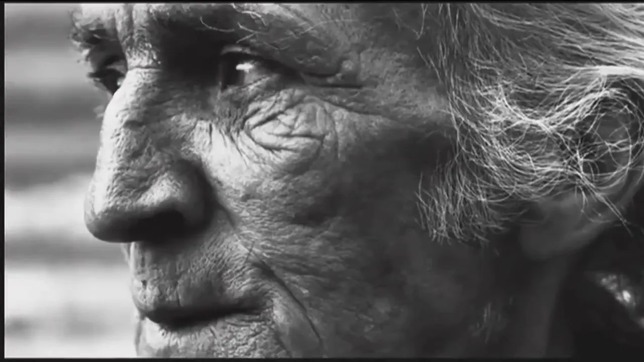 A black and white extreme closeup shows the wrinkled face of an older person with long hair, looking off-camera.