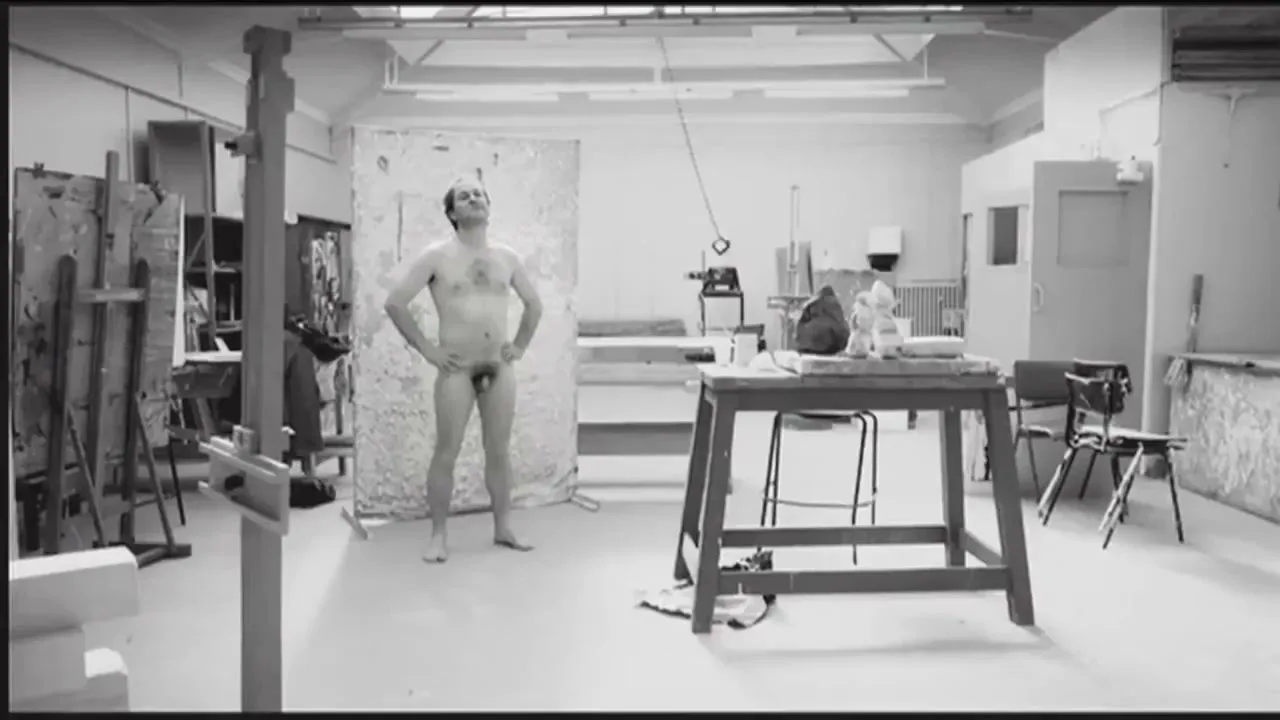 A middle-aged man stands naked with hands on hips, looking up, in a brightly lit art studio with easels and tables.