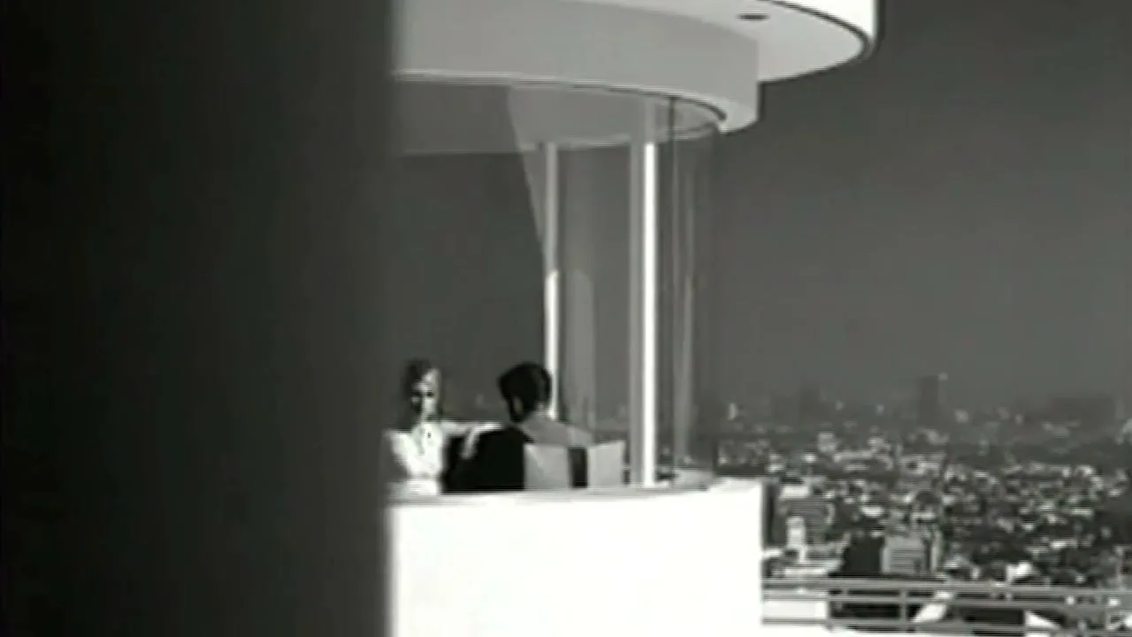 A man and woman sit in a glass-enclosed balcony overlooking a city skyline, captured in black and white.