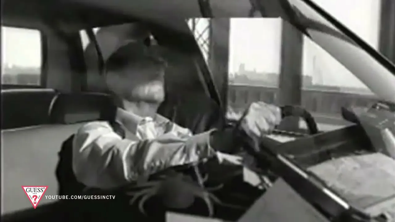 A man in a white shirt and suspenders drives a car, looking forward with a serious expression, in a black and white shot.