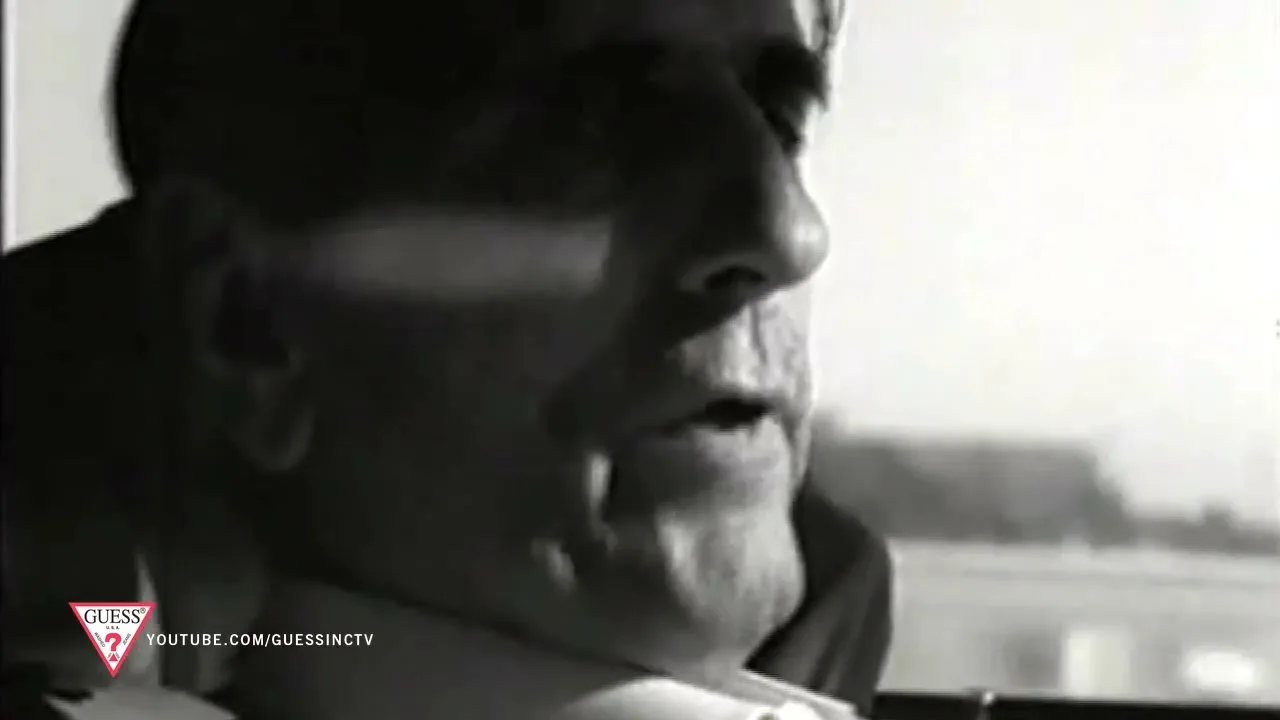 A man with dark hair and a light shirt is seen in profile, looking out a window, in a black and white shot.