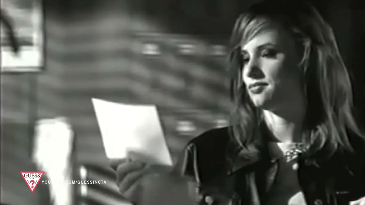 A young woman in a leather jacket holds a piece of paper, looking off-camera with a neutral expression in a dimly lit interior.