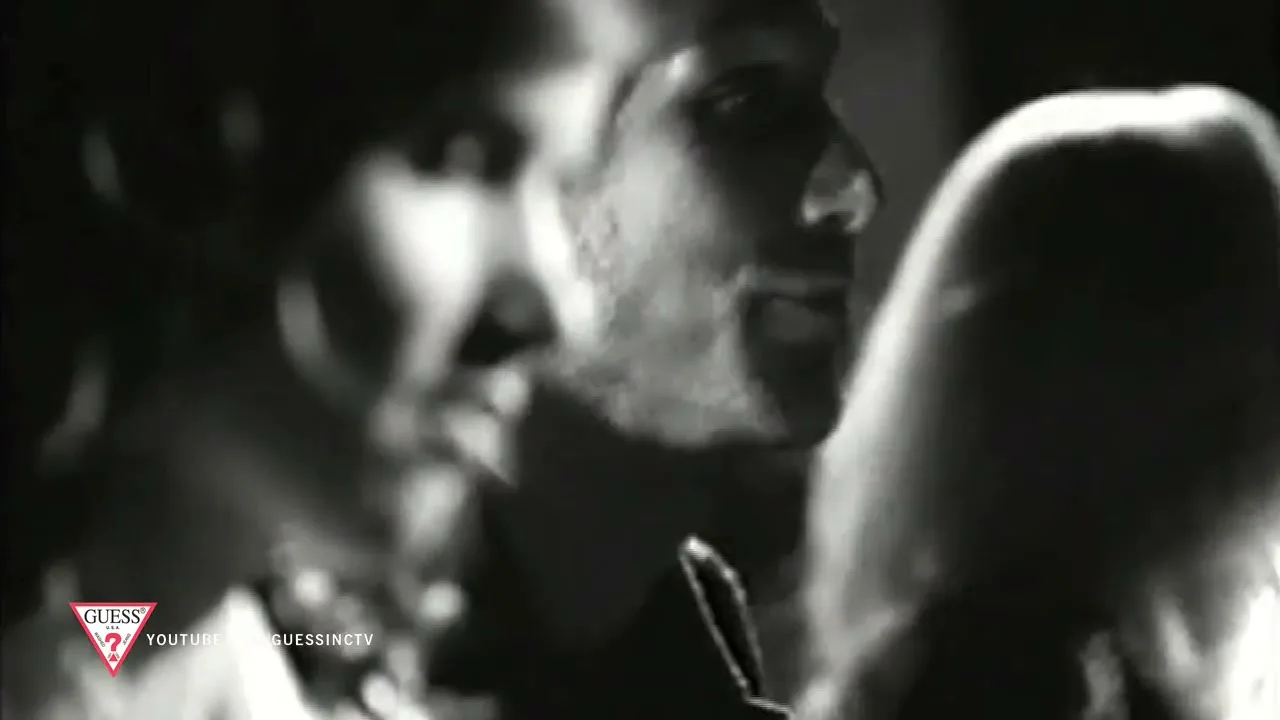 A man and woman are seen in a medium closeup, with the man's face in profile, as they appear to be whispering to each other in a dark, intimate setting.
