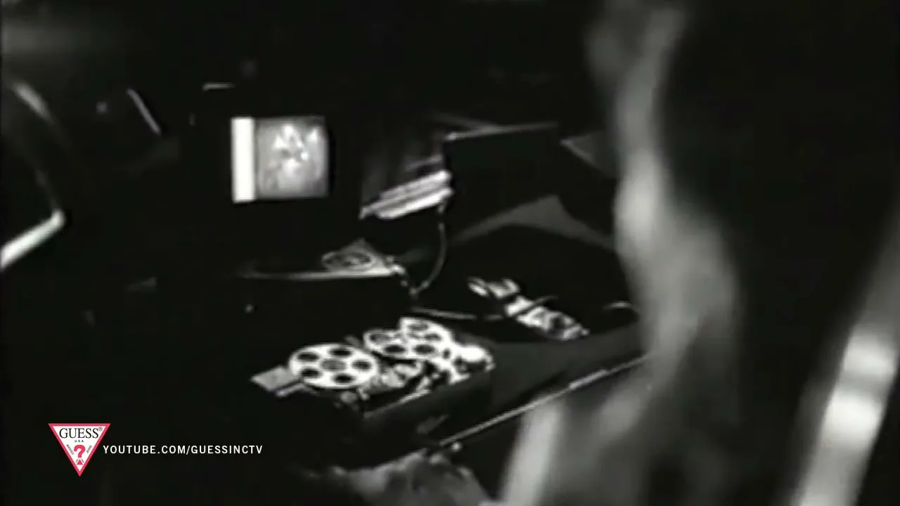 A person's head and shoulder are visible in the foreground, looking at a small monitor and film reels on a desk in a dark room.