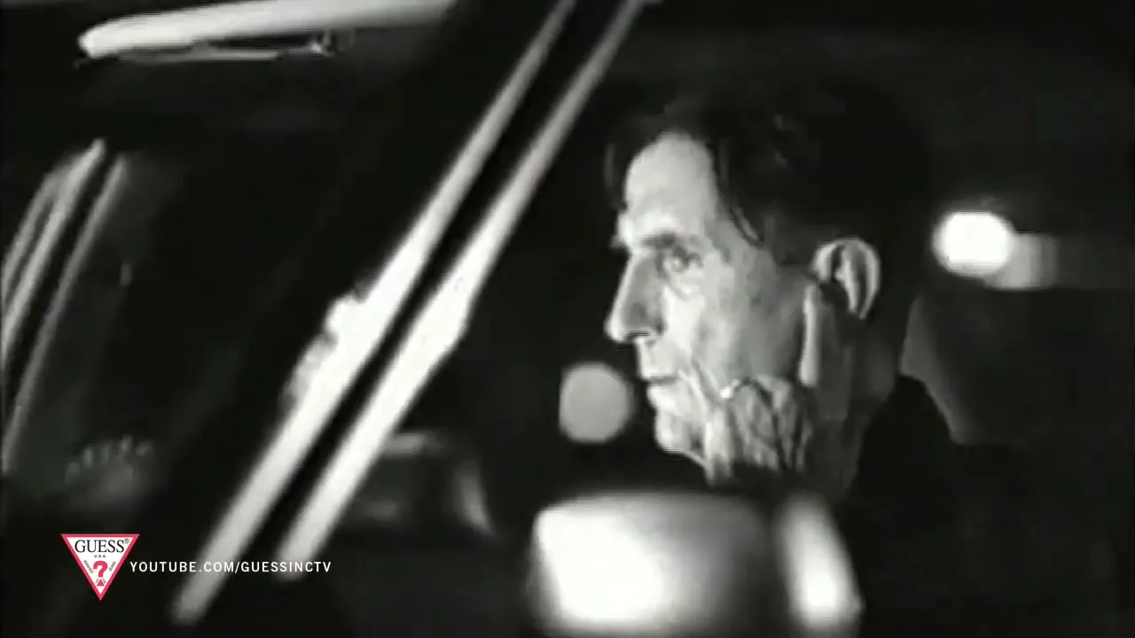 A man with dark hair and a striped shirt drives a car at night, looking forward with a serious expression.