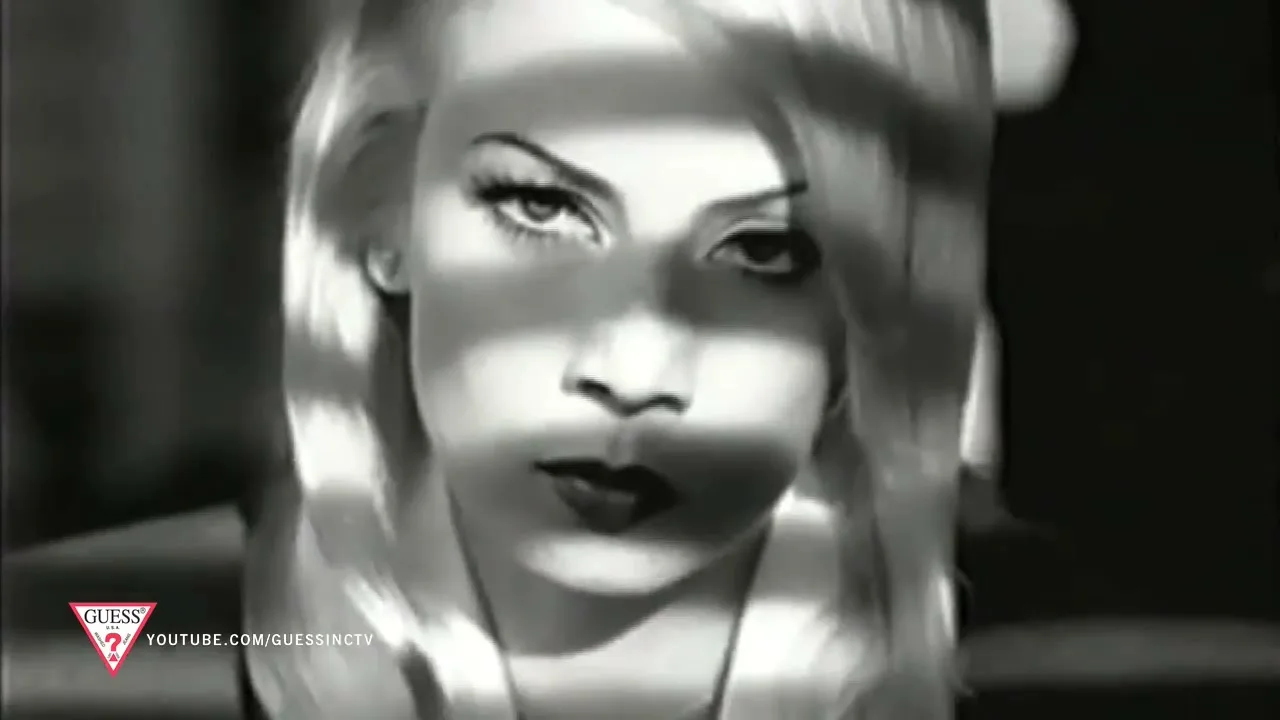 A blonde woman with heavy makeup looks up at the camera, with shadows cast across her face, in a black and white shot.