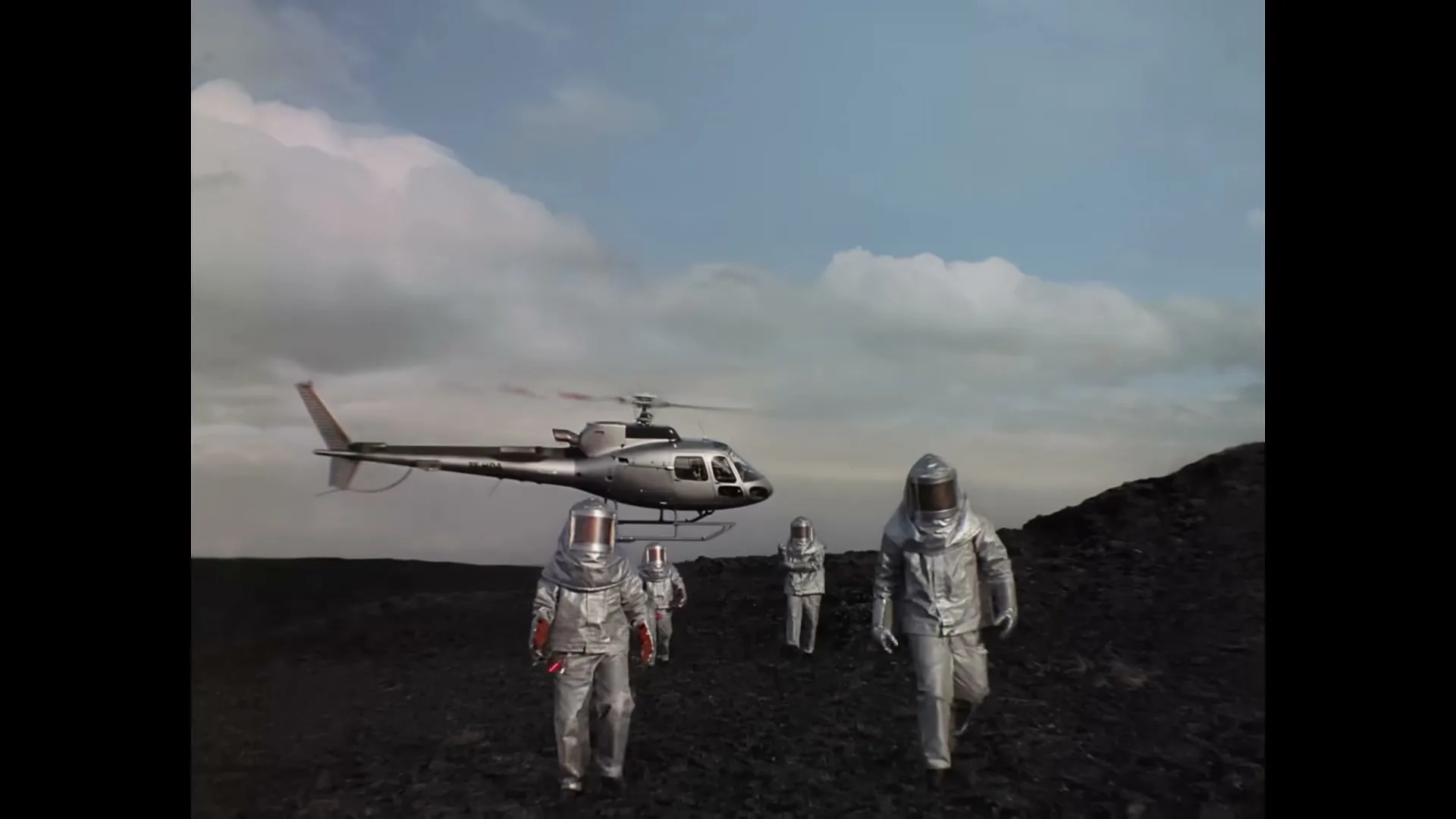 A group of people in silver hazmat suits walk away from a helicopter on a dark, rocky landscape under a cloudy sky.