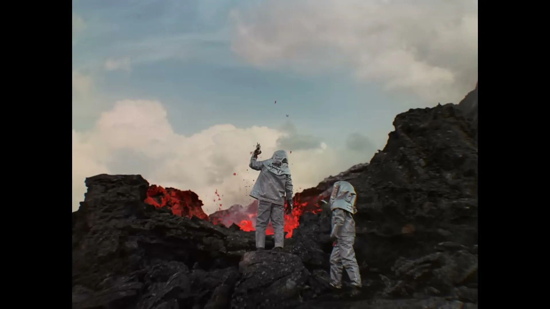 Two people in silver suits stand on a rocky volcanic landscape, one throwing something red into the air, with lava glowing in the background under a cloudy sky.