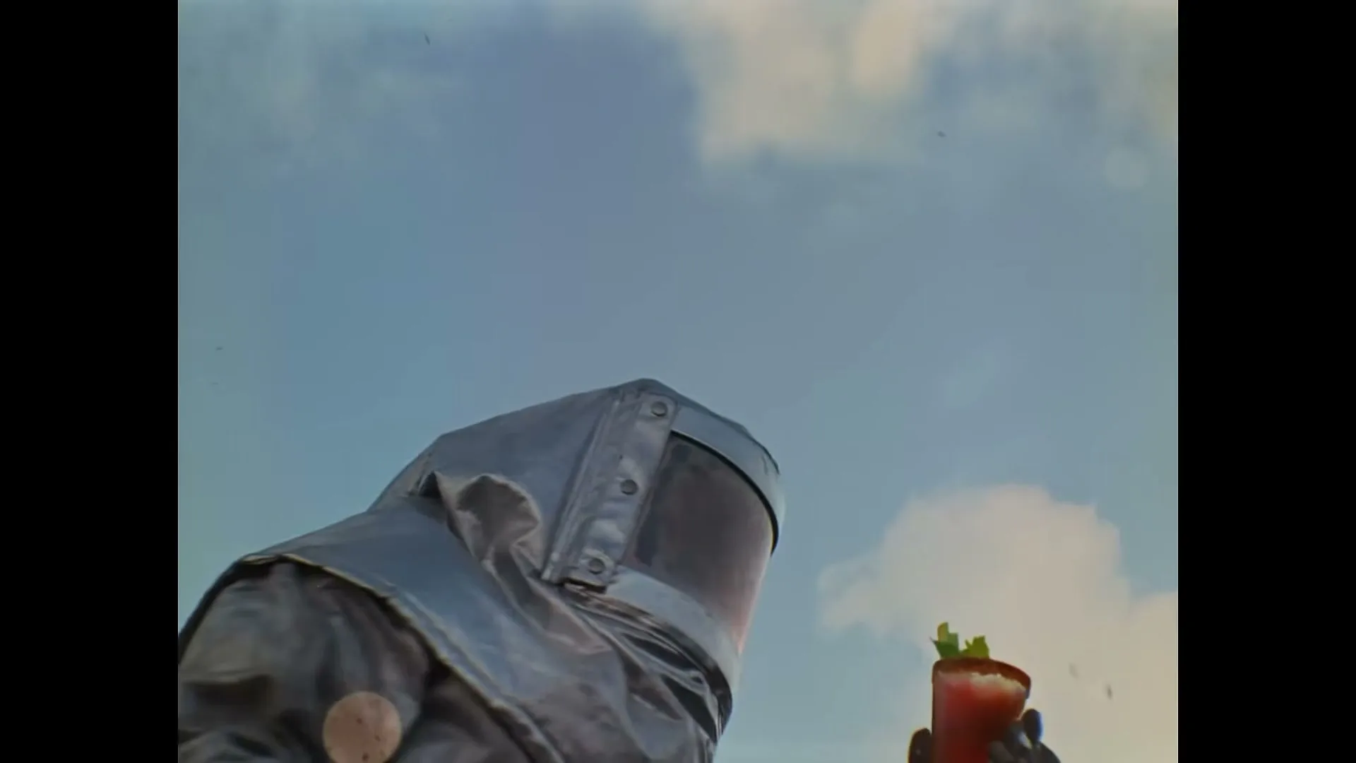 A person in a silver hazmat suit holds a Bloody Mary cocktail as red liquid splashes around them under a blue sky.