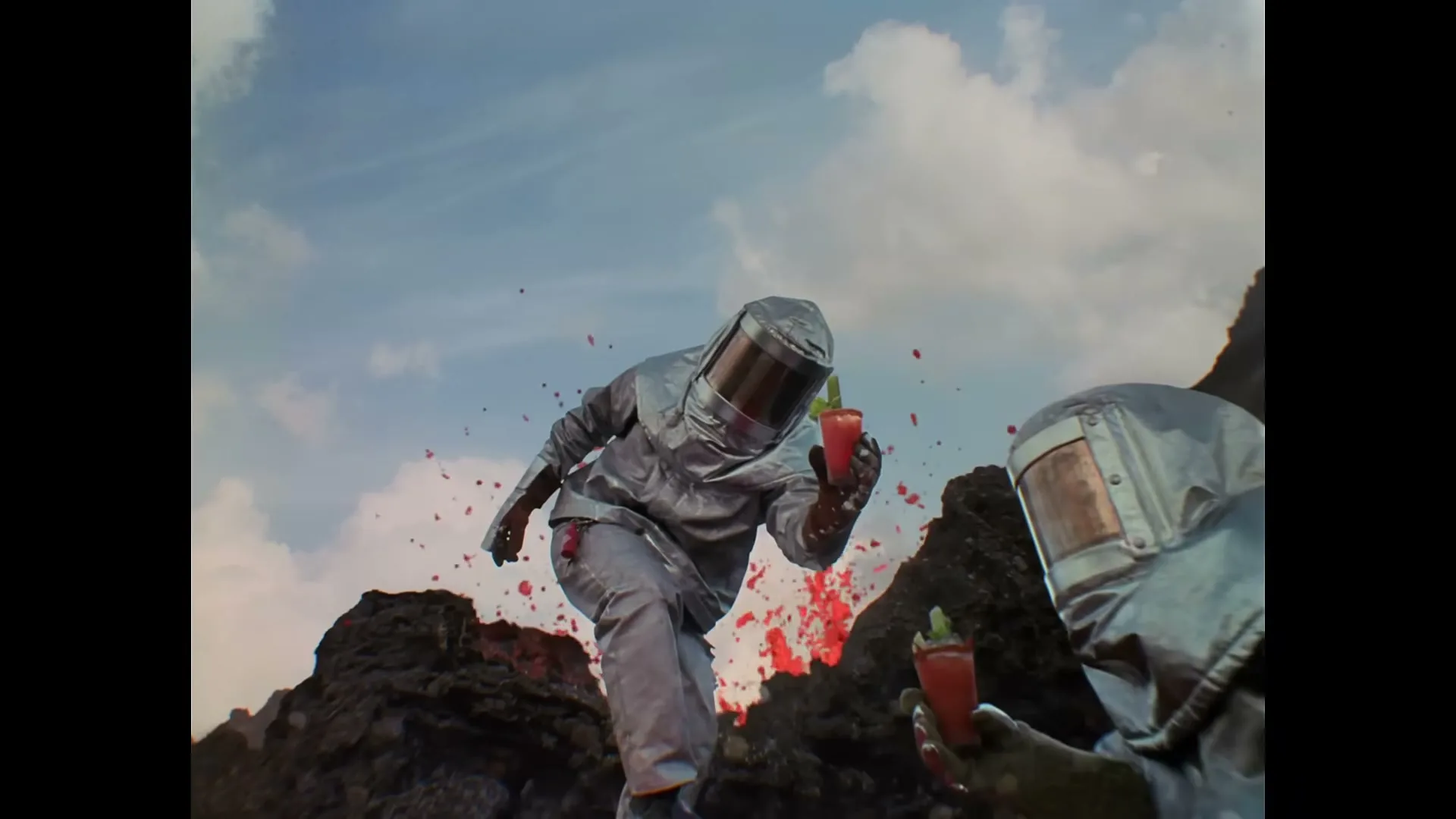 Two people in silver hazmat suits hold red cocktails, with red liquid splashing around them on a rocky, alien landscape under a cloudy sky.