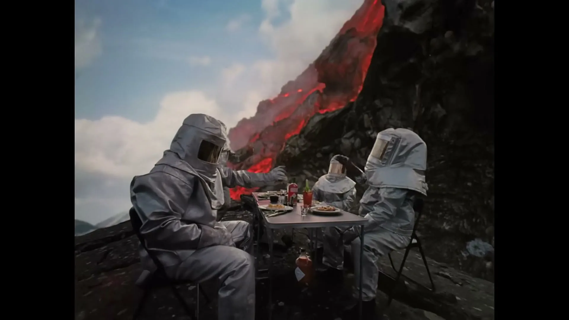 Four people in silver heat-resistant suits are having a meal at a table on a rocky cliff, with a river of lava flowing down a mountain in the background under a blue sky.