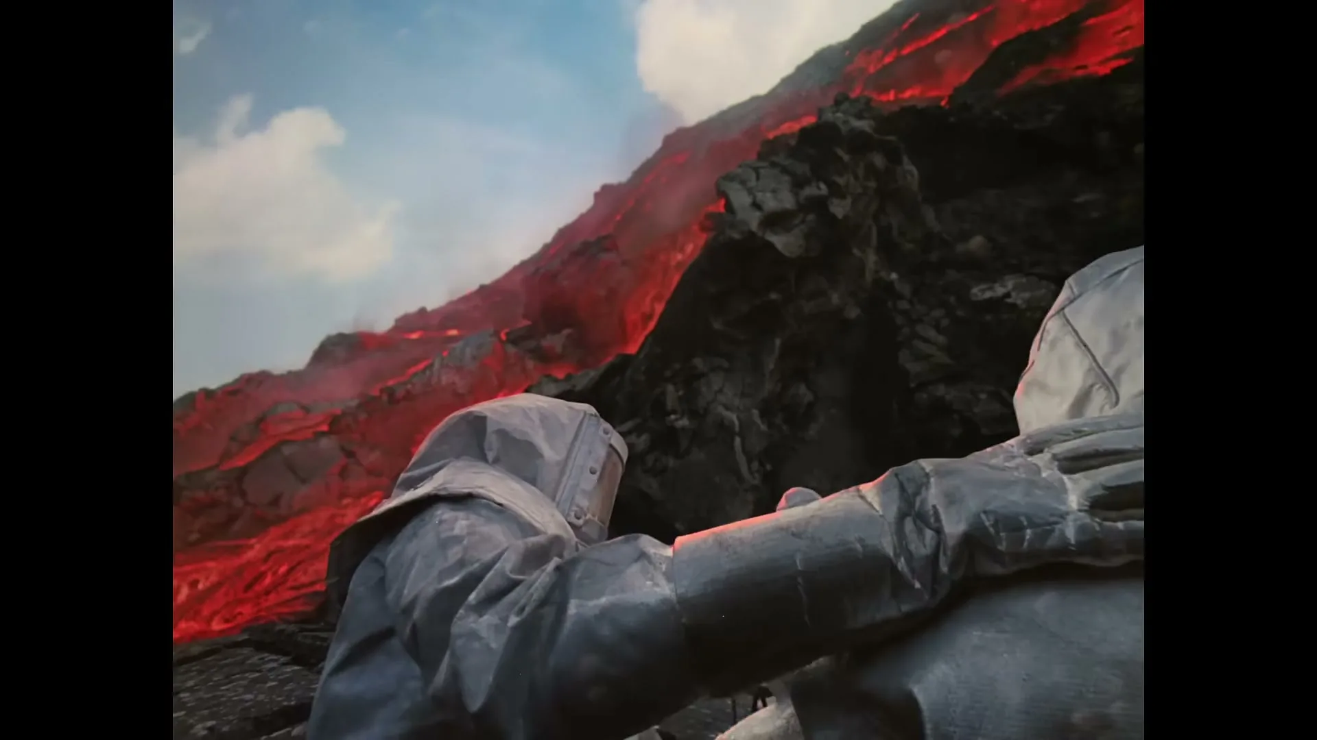Two people in grey hazmat suits are seen from behind, with one person's arm around the other, as a lava flow cascades down a mountain in the background.