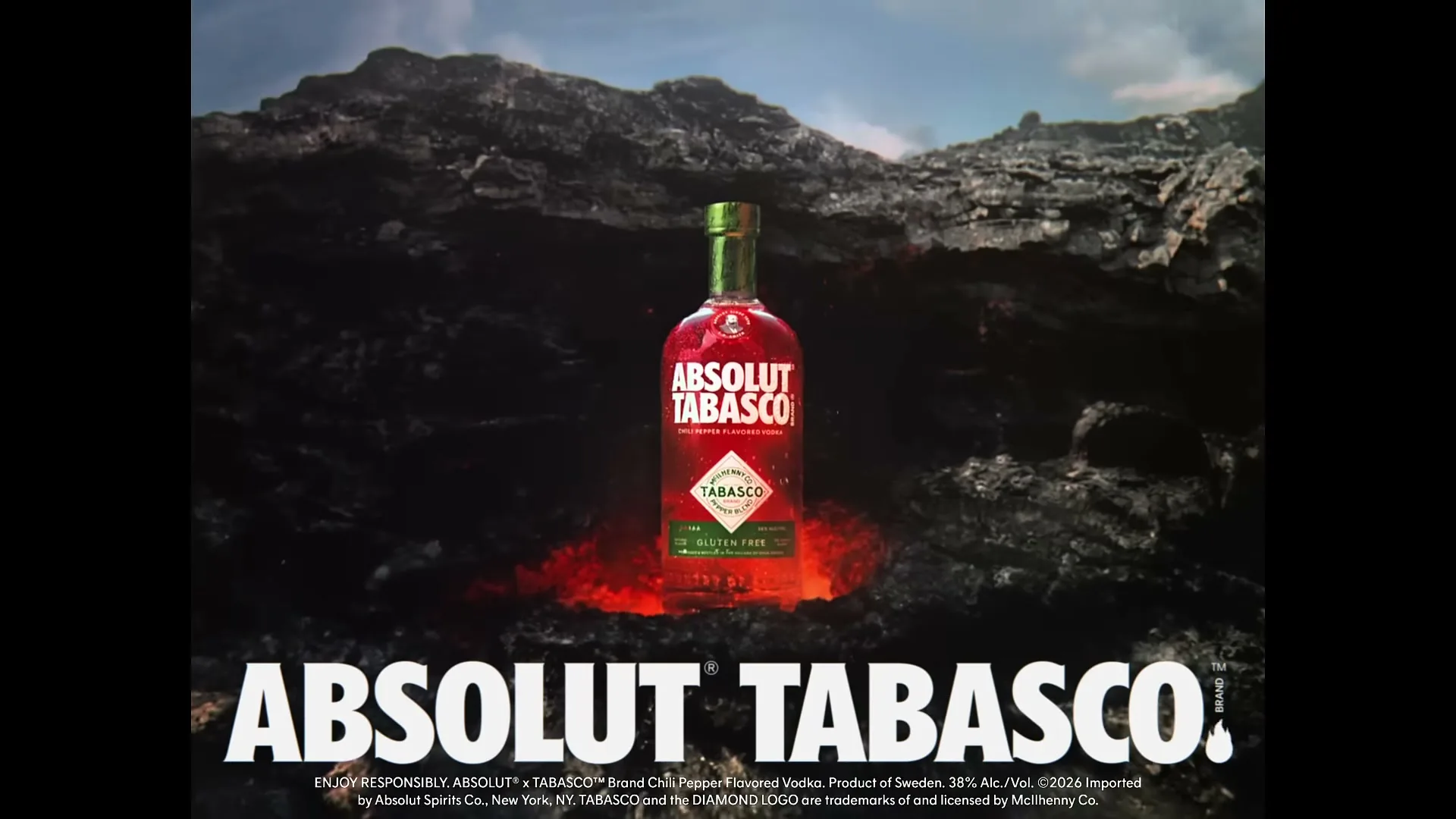 A bottle of Absolut Tabasco vodka stands on a bed of glowing red embers amidst a dark, rocky landscape under a cloudy sky.