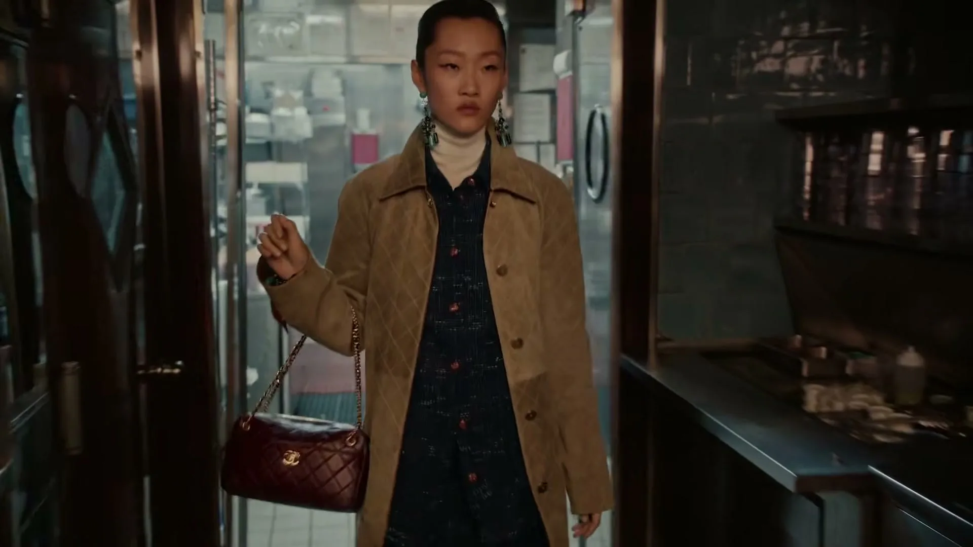 A female model in a quilted coat and carrying a handbag walks through a restaurant kitchen, looking around with a neutral expression.