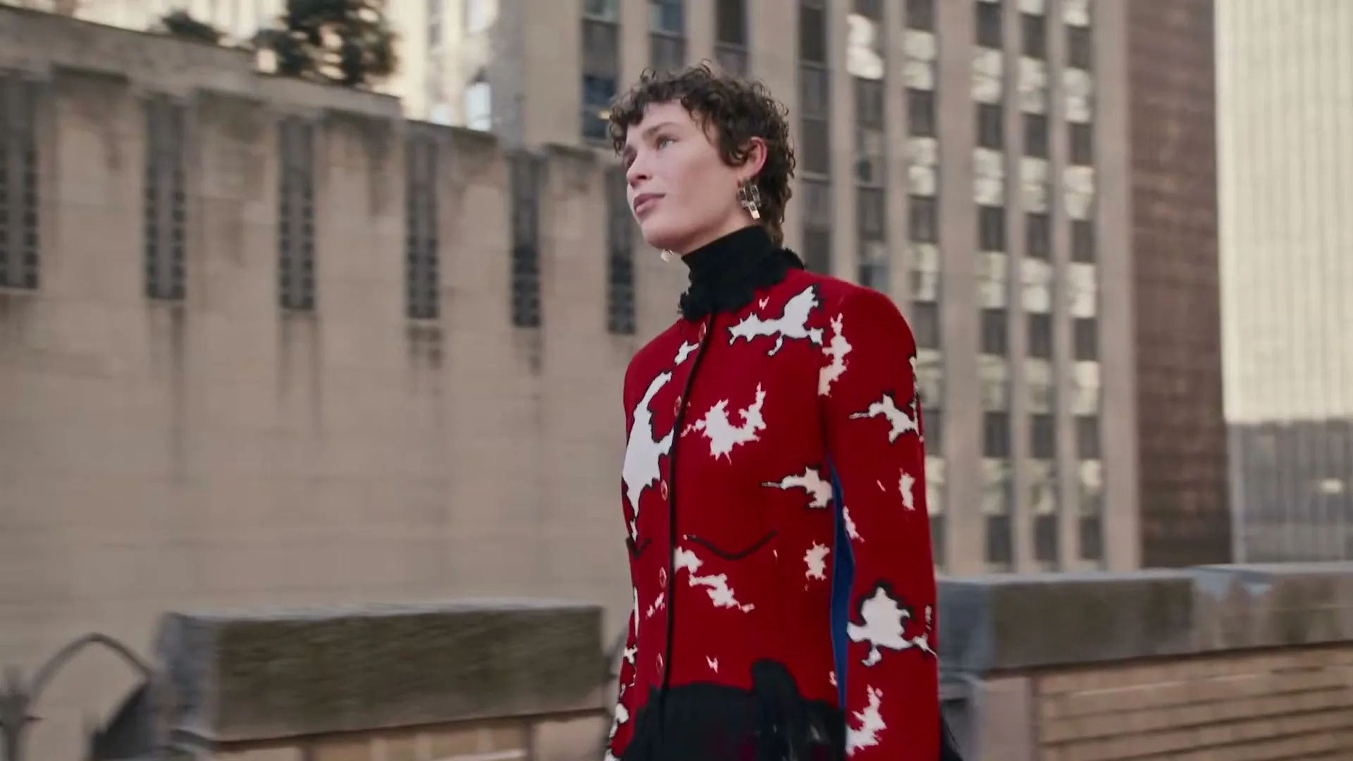 A female model with short curly hair walks on a rooftop in a red and white patterned jacket, looking up and to the left.
