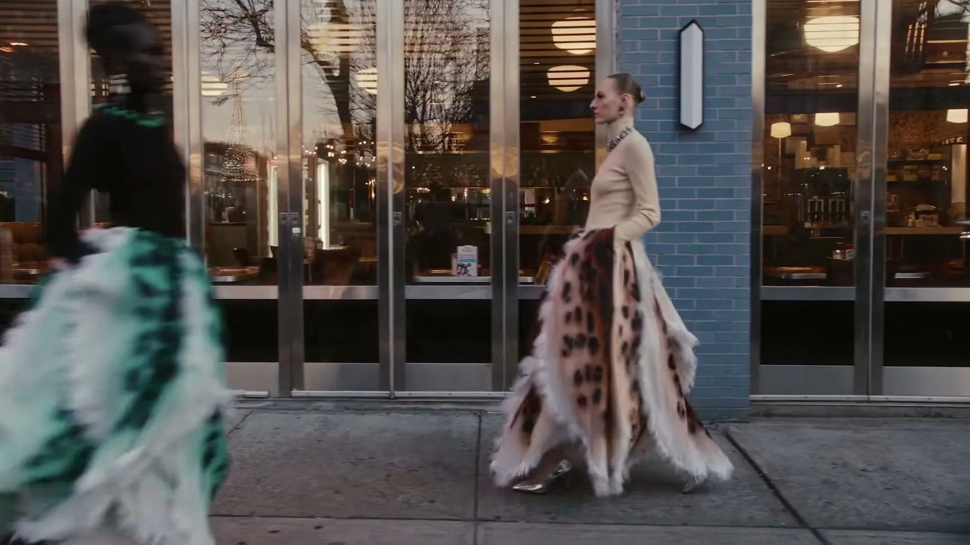 A female model in a beige top and a long, spotted, feathery skirt walks past a storefront with another model in a green and white skirt passing in the foreground.