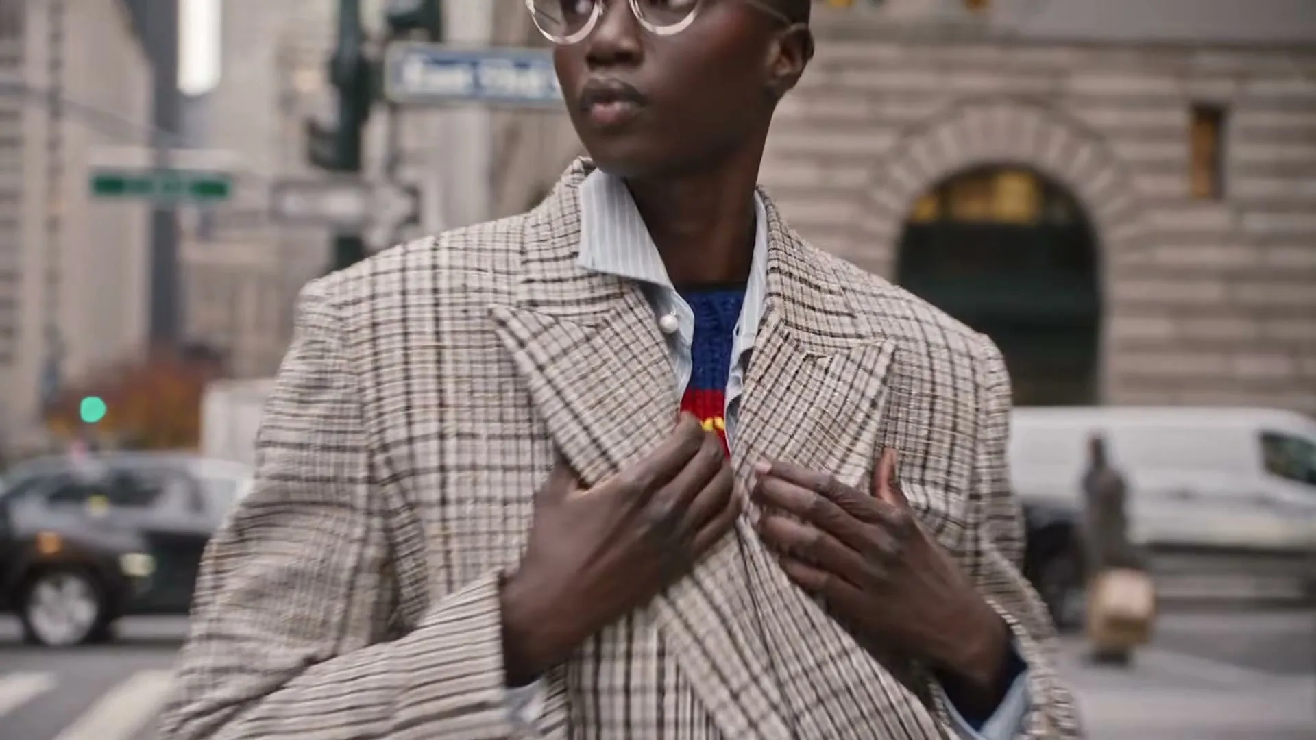A dark-skinned model with a shaved head and glasses walks on a city sidewalk, revealing a Superman sweater under her plaid jacket.
