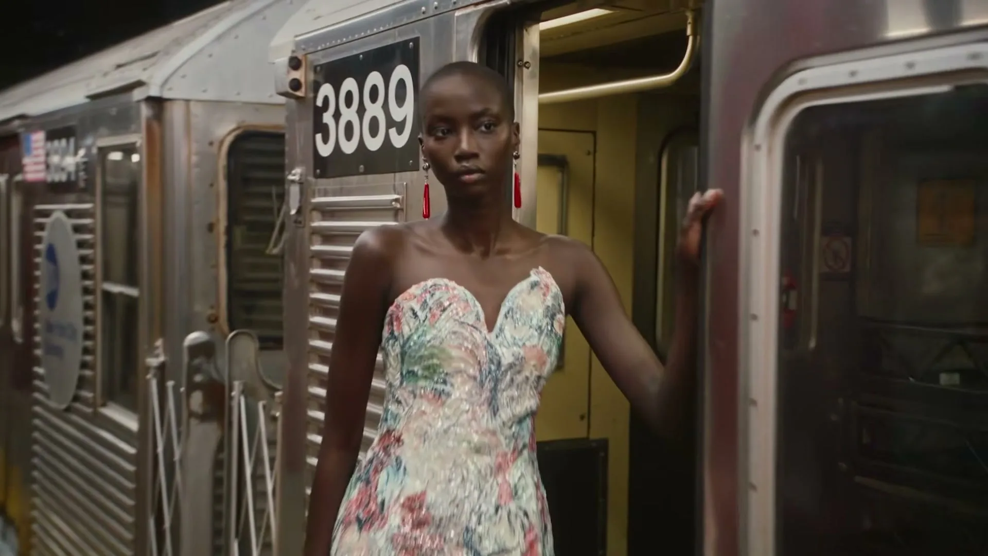 A dark-skinned model with a shaved head and a floral dress stands in a subway car doorway, looking to the side.