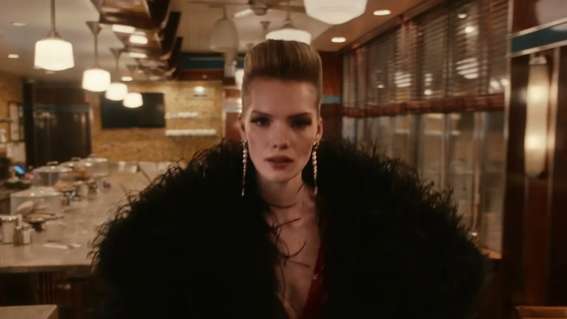 A female model with a dramatic hairstyle and a black feathered coat walks through a retro diner, looking around with a serious expression.
