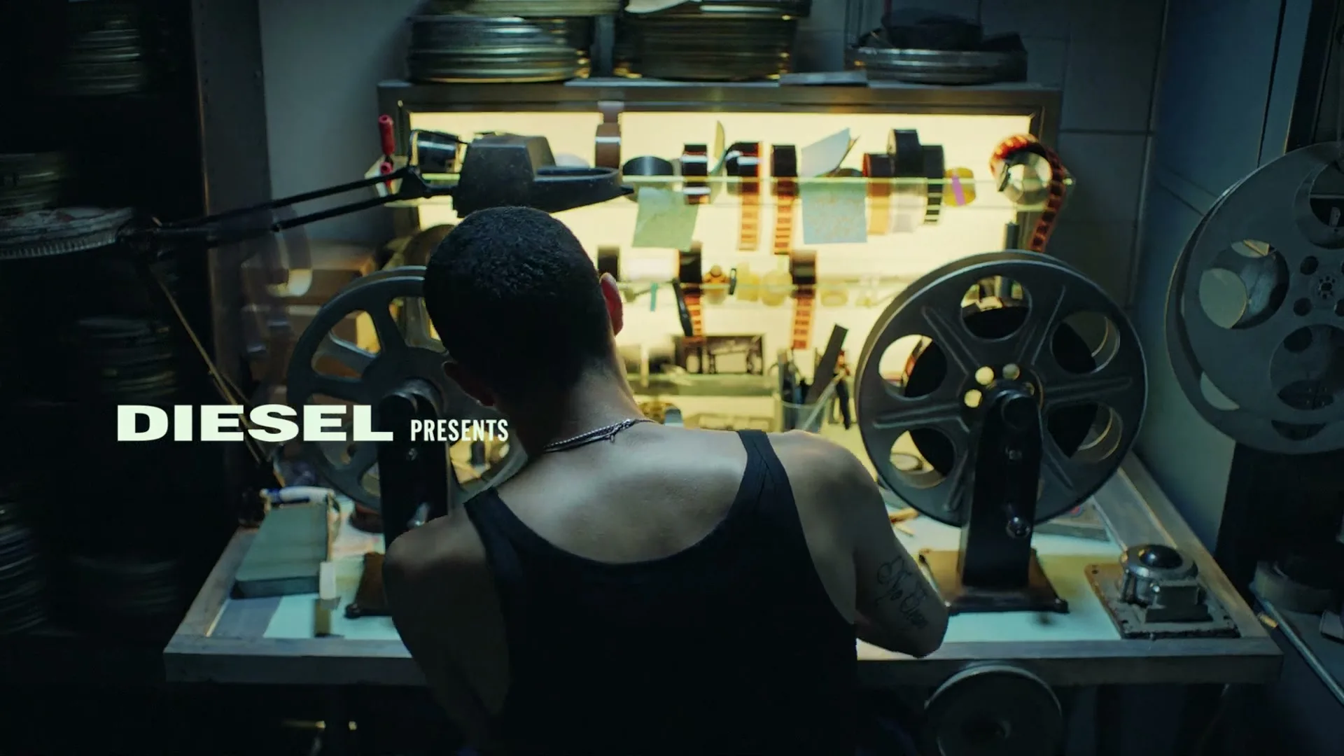 A young man with short dark hair and a black tank top sits with his back to the camera, working at a film editing machine in a dimly lit room.
