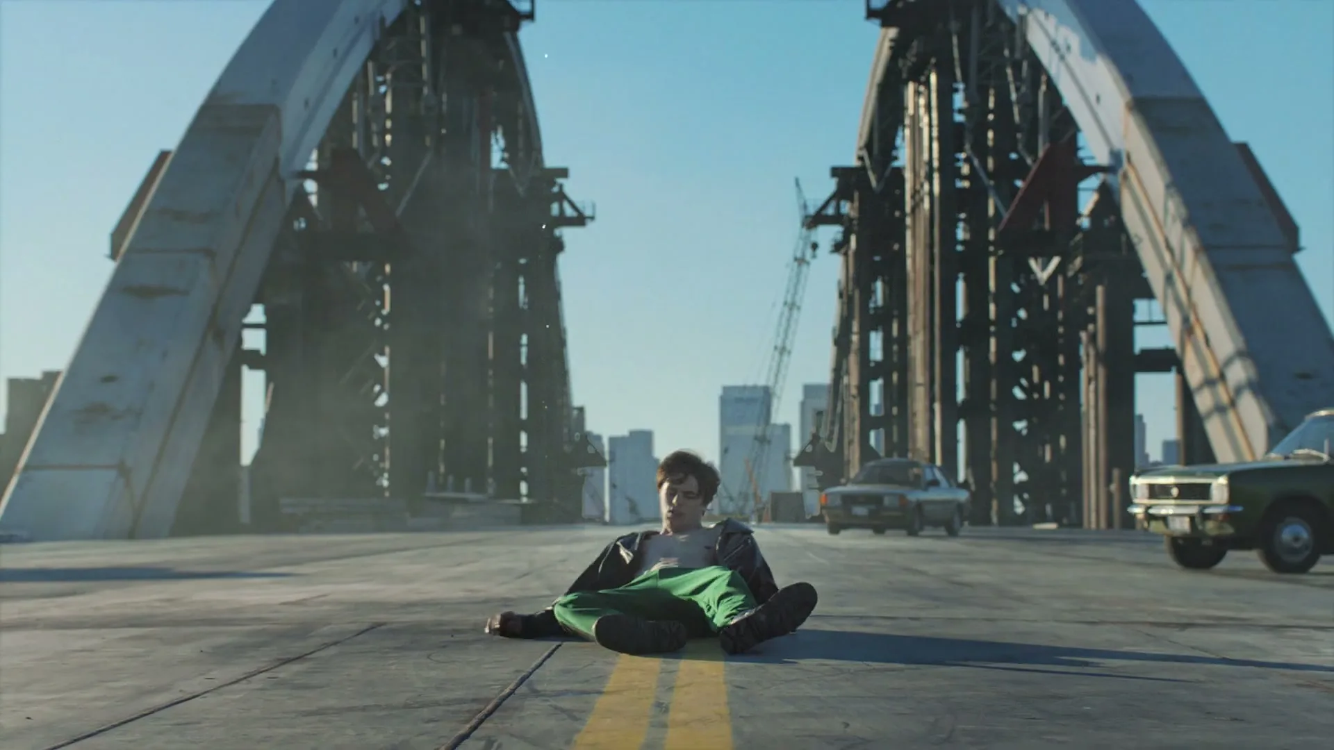 A young man in green pants and a black jacket sits on a bridge, looking up at the sky with a city skyline in the background.