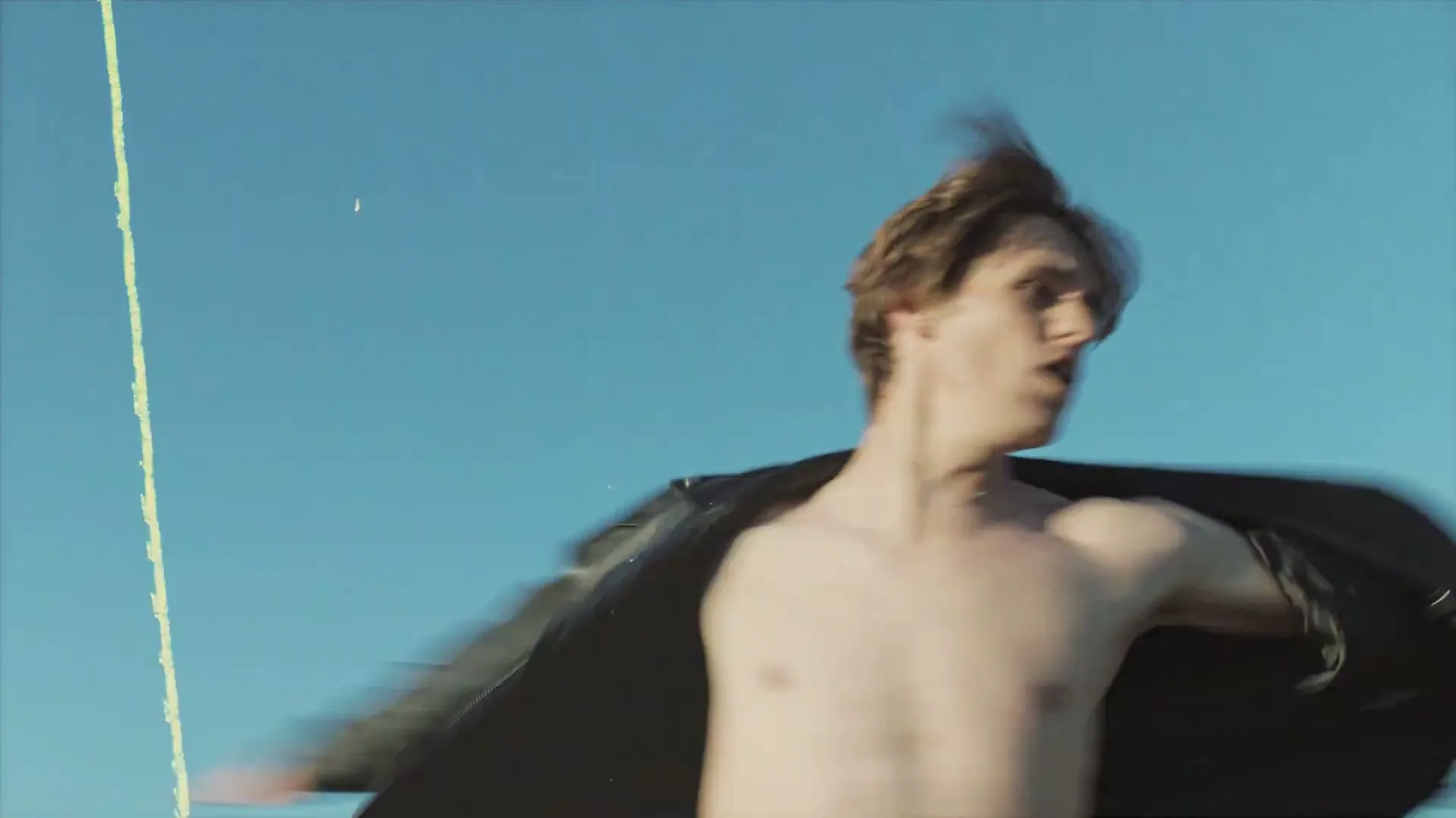 A young man with brown hair and a black leather jacket spins around against a clear blue sky.
